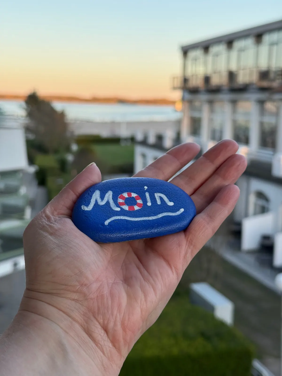 Bemalter Stein mit der Aufschrift Moin in einer Hand, dahinter der Blick vom Balkon auf die Ostsee