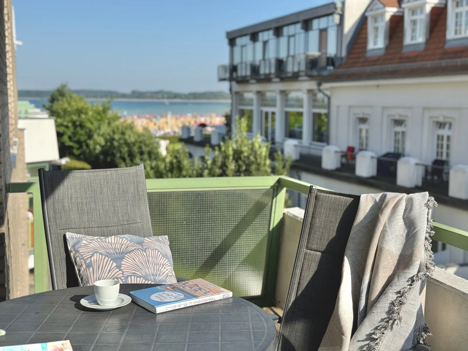 Südbalkon mit rundem Tisch, Kaffeetasse und Buch, dahinter der Blick auf den Kurstrand und die Ostsee
